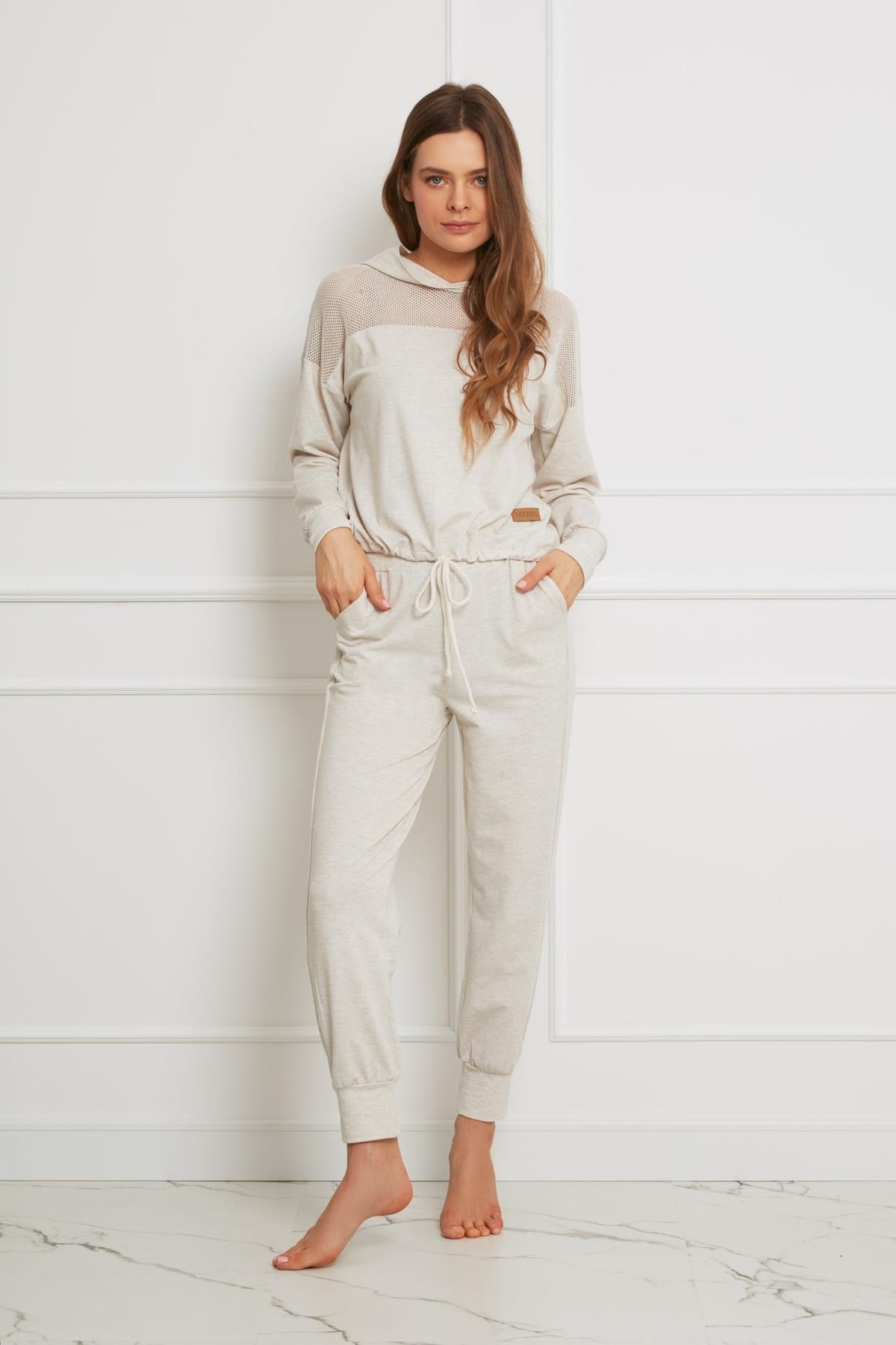 Kobieta z długimi brązowymi włosami stoi boso w pomieszczeniu, ubrana w Loungewear wiskozowe spodnie homewear - beżowy melanż i pasującą bluzę z kapturem, z rękami w kieszeniach, na tle białej ściany wyłożonej panelami.