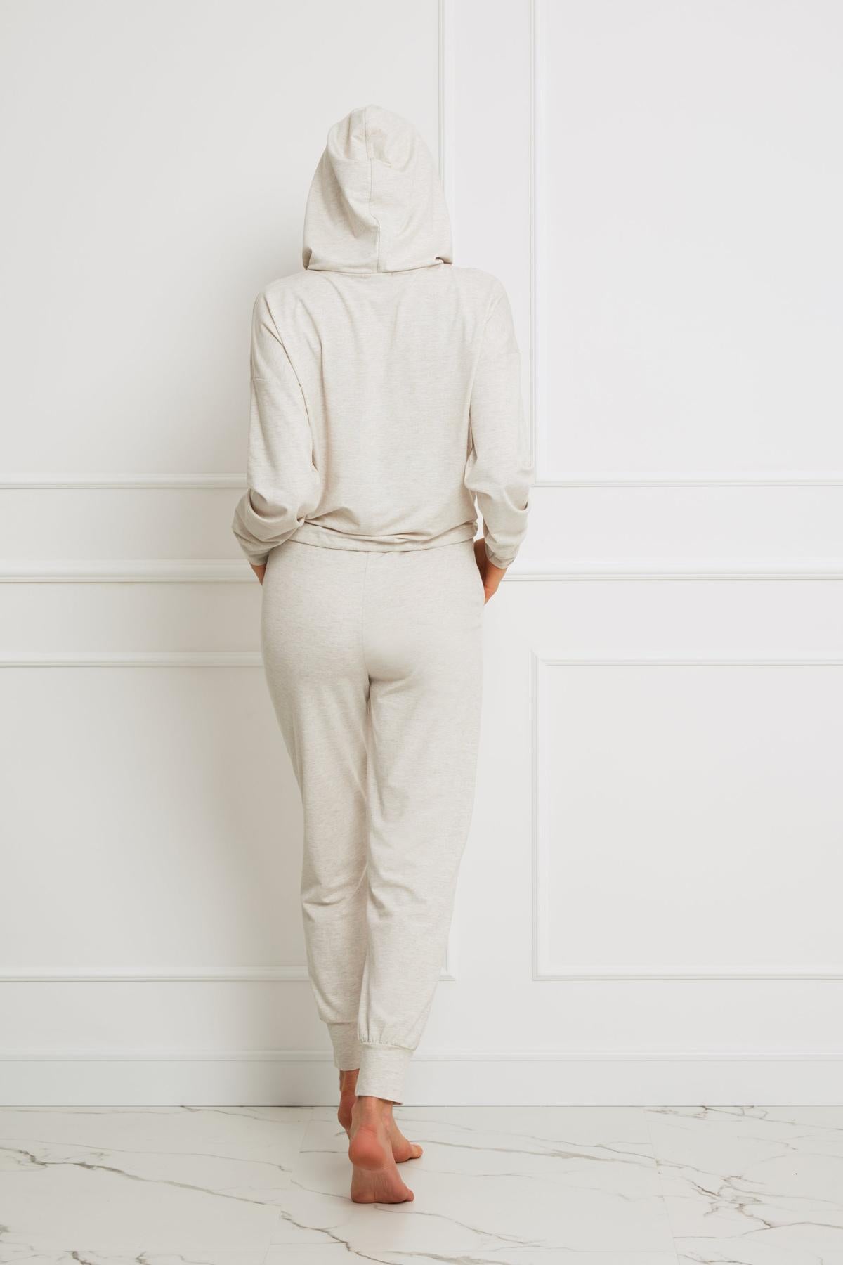 Osoba stoi boso twarzą do białej ściany, ręce w kieszeniach, ubrana w beżowy melanż Loungewear wiskozowe spodnie homewear z dopasowaną bluzą z kapturem i naciągniętym kapturem.