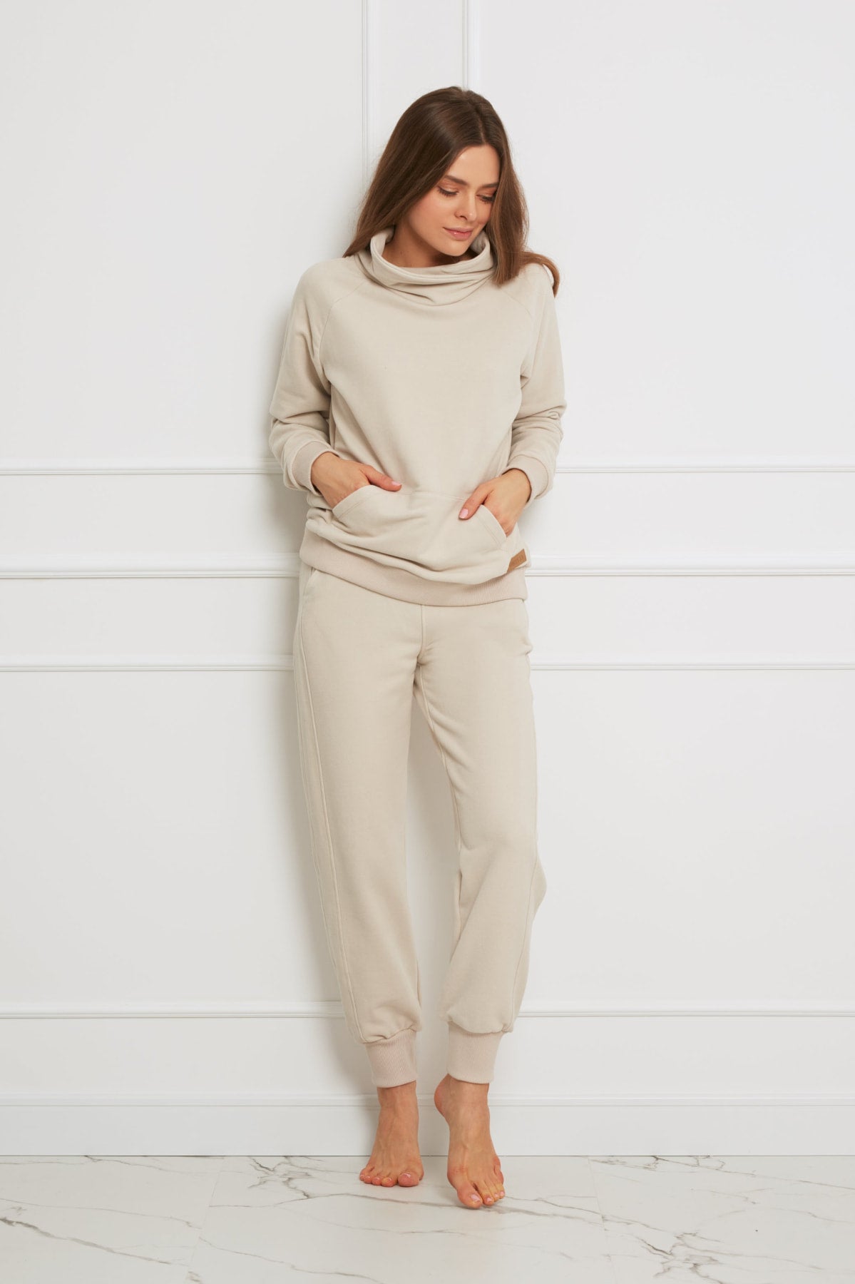 Kobieta stoi boso przy białej ścianie, ubrana w bluza bawełniana Loungewear z wywijanym kołnierzem - beżowa z pasującymi spodniami dresowymi jogger. Jej ręce są w kieszeniach bluzy, gdy patrzy w dół.