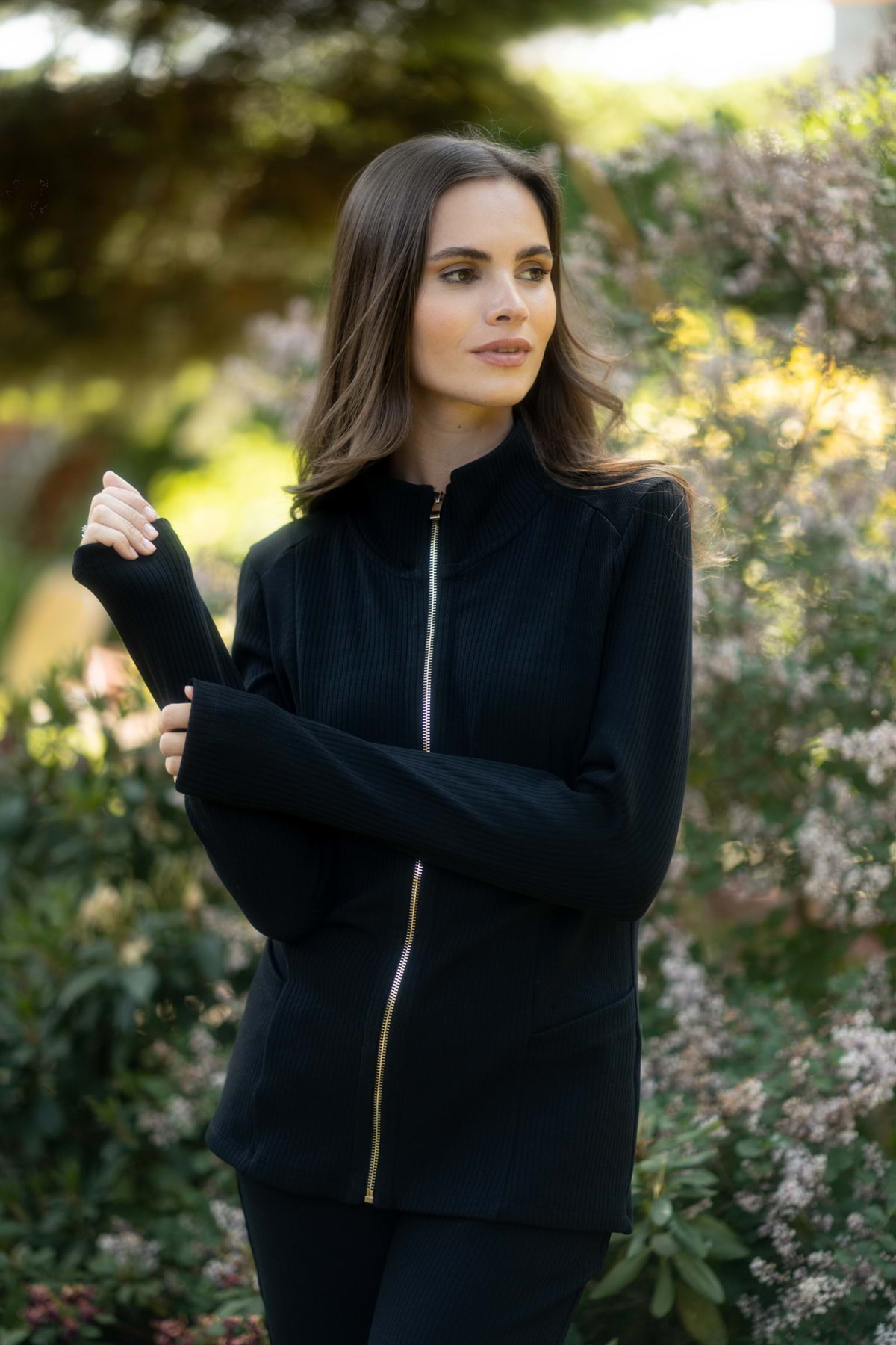 Kobieta z długimi brązowymi włosami stoi na zewnątrz wśród zielonych krzewów i kwiatów, ubrana w Loungewear bluza ze stójką i suwakiem - czarna i dopasowane spodnie, patrząc w bok z podniesioną ręką.