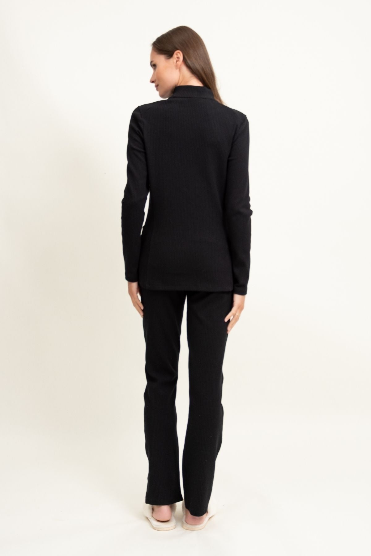 Kobieta z prostymi brązowymi włosami, ubrana w Loungewear bluza ze stójką i suwakiem - czarna i czarne spodnie z jasnymi butami, na białym tle.