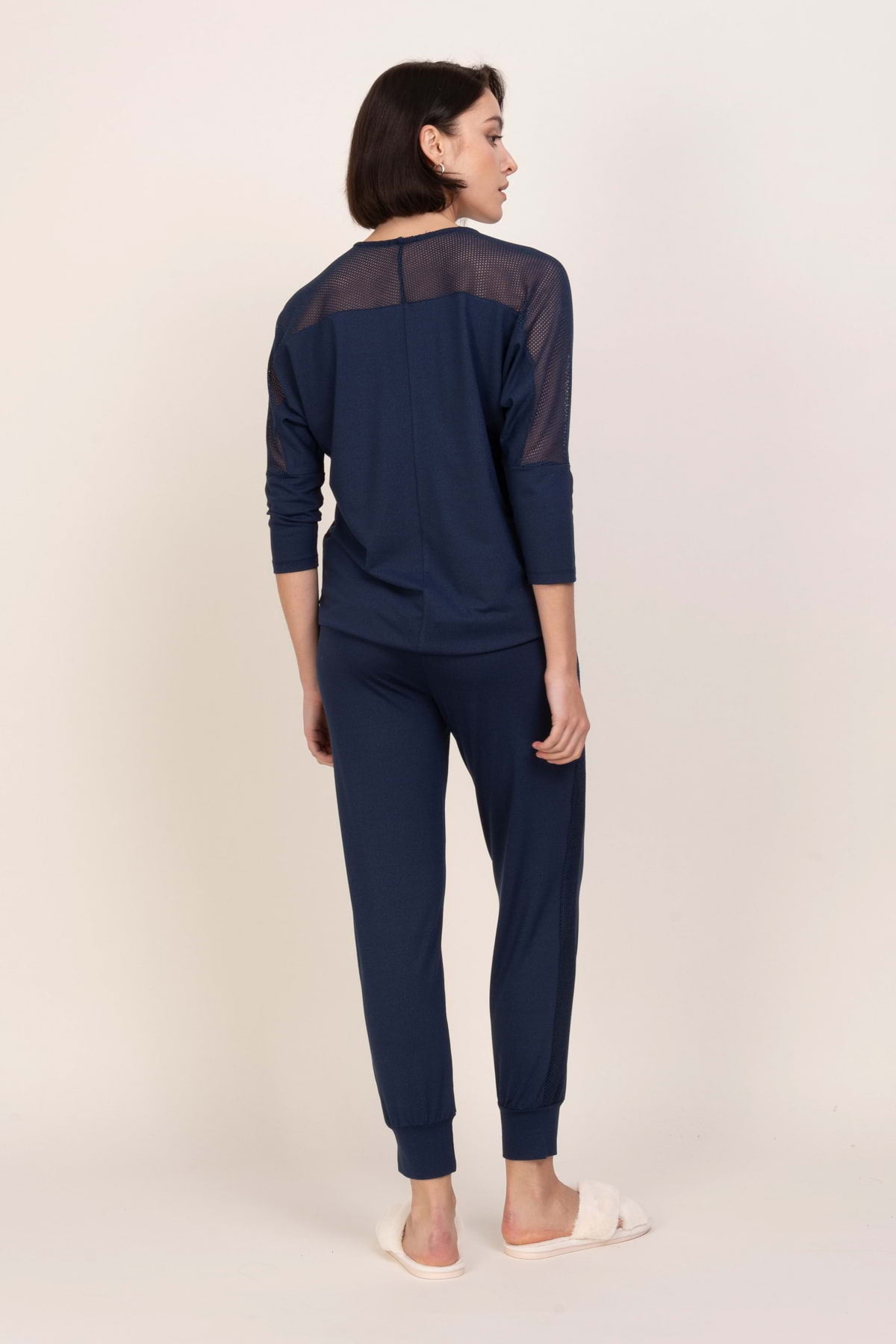 Kobieta stoi odwrócona tyłem, ubrana w zestaw Loungewear wiskozowa bluzka z siateczką - ciemny dżins z siateczkowymi detalami w górnej części pleców i pasującymi spodniami. Ma na sobie białe pantofle na jasnym tle.