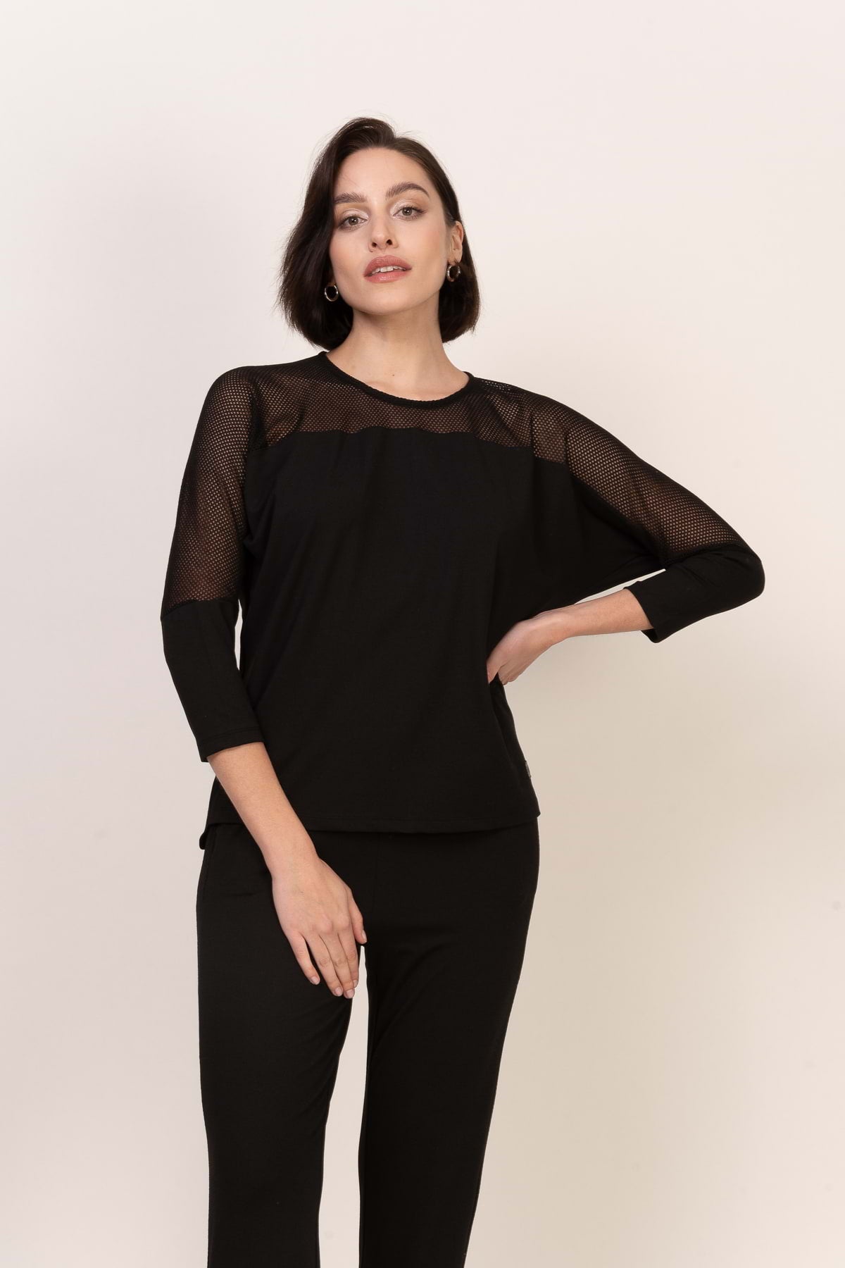 Kobieta z krótkimi ciemnymi włosami pozuje w Loungewear wiskozowa bluzka z siateczką - czarna z kimonowym krojem i rękawami trzy czwarte, stojąc na jasnym tle z jedną ręką na biodrze.