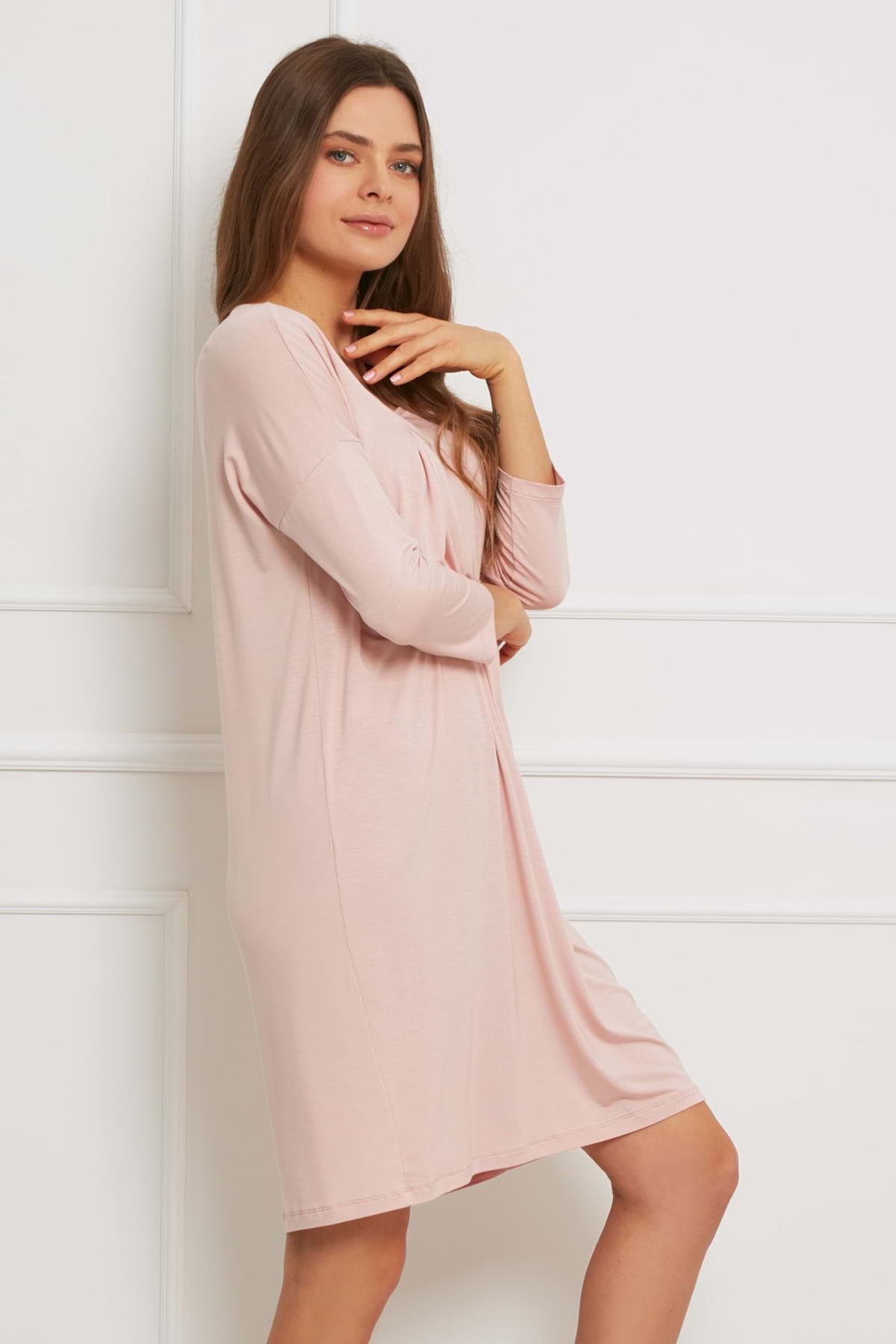 Kobieta z długimi brązowymi włosami pozuje na tle białej ściany, delikatnie uśmiechnięta i ubrana w Loungewear wiskozowa luźna sukienka - brudny róż, jej dłoń spoczywa w pobliżu podbródka.