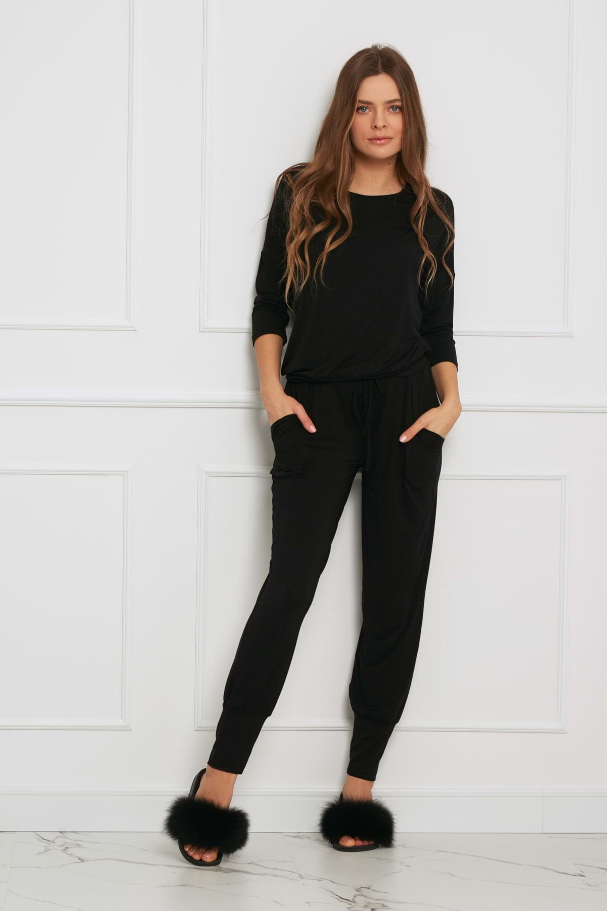 Kobieta o długich brązowych włosach nosi Loungewear długi komplet ze spodniami z kieszeniami - czarny, łącząc go z czarnymi futrzanymi kapciami, stojąc przy białej ścianie wyłożonej panelami, z rękami w kieszeniach i jedną nogą lekko ugiętą.