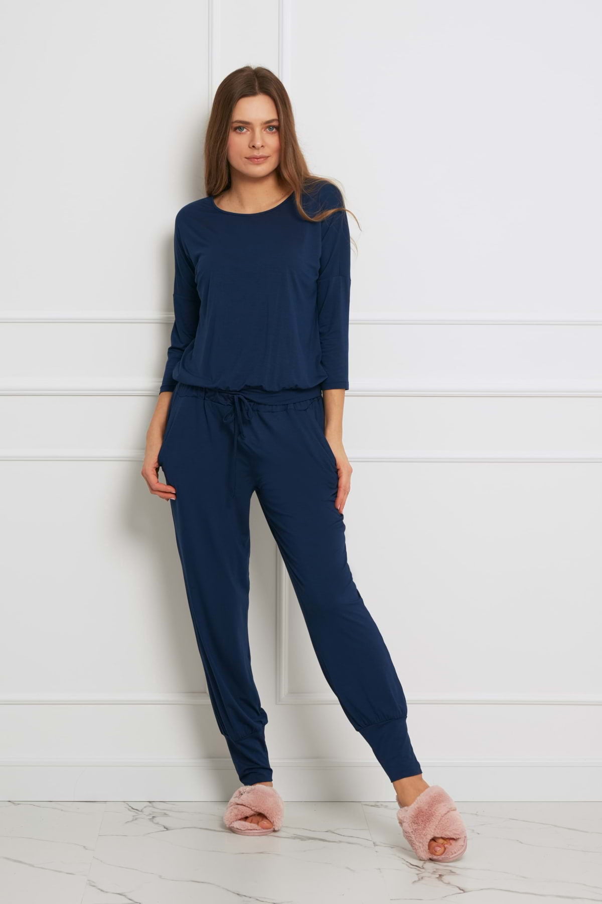 Kobieta stoi pod białą ścianą w Loungewear długi komplet ze spodniami z kieszeniami - granatowy, w połączeniu z jasnoróżowymi rozmytymi kapciami. Jej długie brązowe włosy są rozpuszczone i wygląda na zrelaksowaną.
