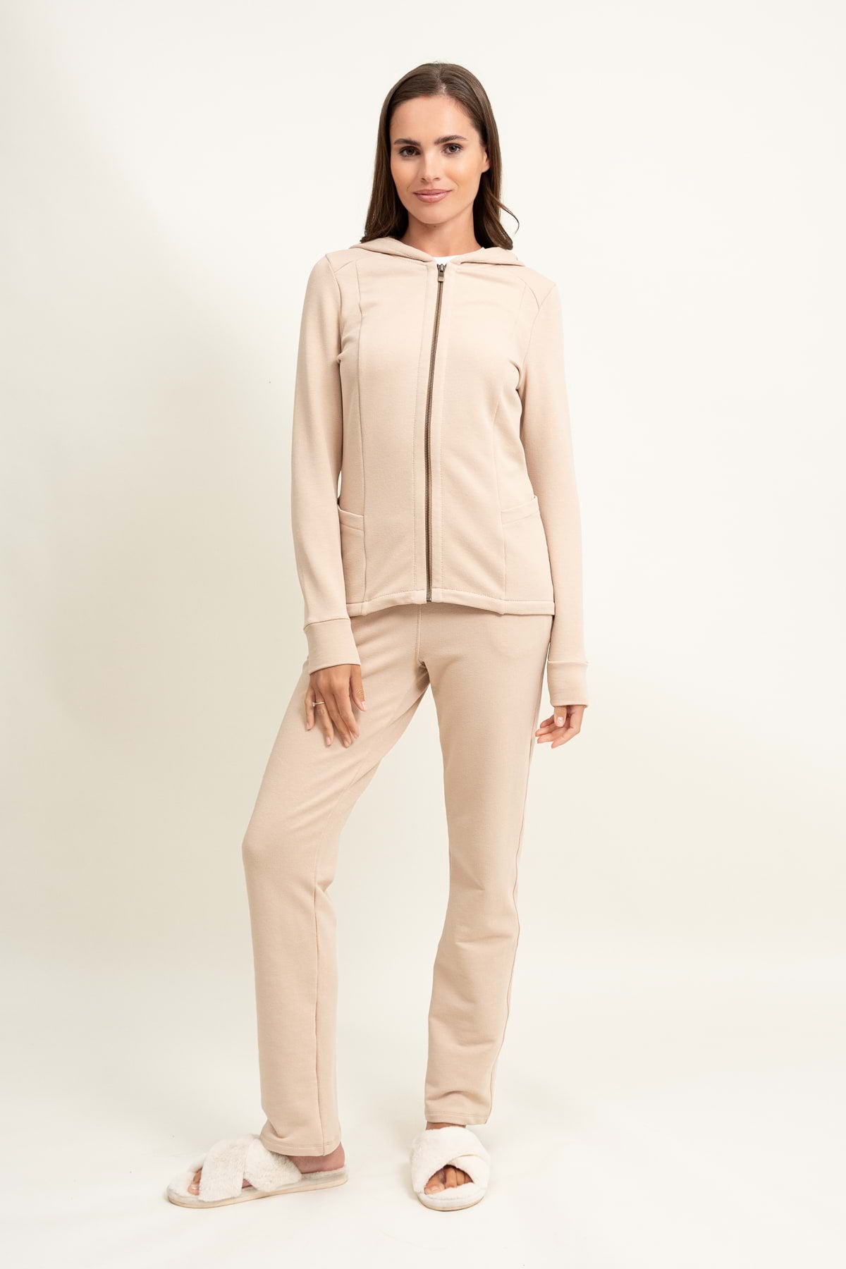 Kobieta z prostymi brązowymi włosami i zrelaksowanym wyrazem twarzy nosi Loungewear rozpinana bluza z kapturem - jasny beż z dopasowanymi spodniami, stojąc na prostym jasnym tle i łącząc strój z białymi rozmytymi kapciami.