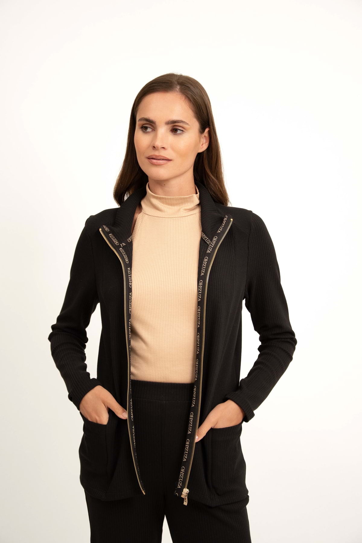 Kobieta z długimi brązowymi włosami, ubrana w beżowy golf i Loungewear bluza ze stójką i suwakiem - czarna z rękami w kieszeniach kurtki, stoi na prostym białym tle.