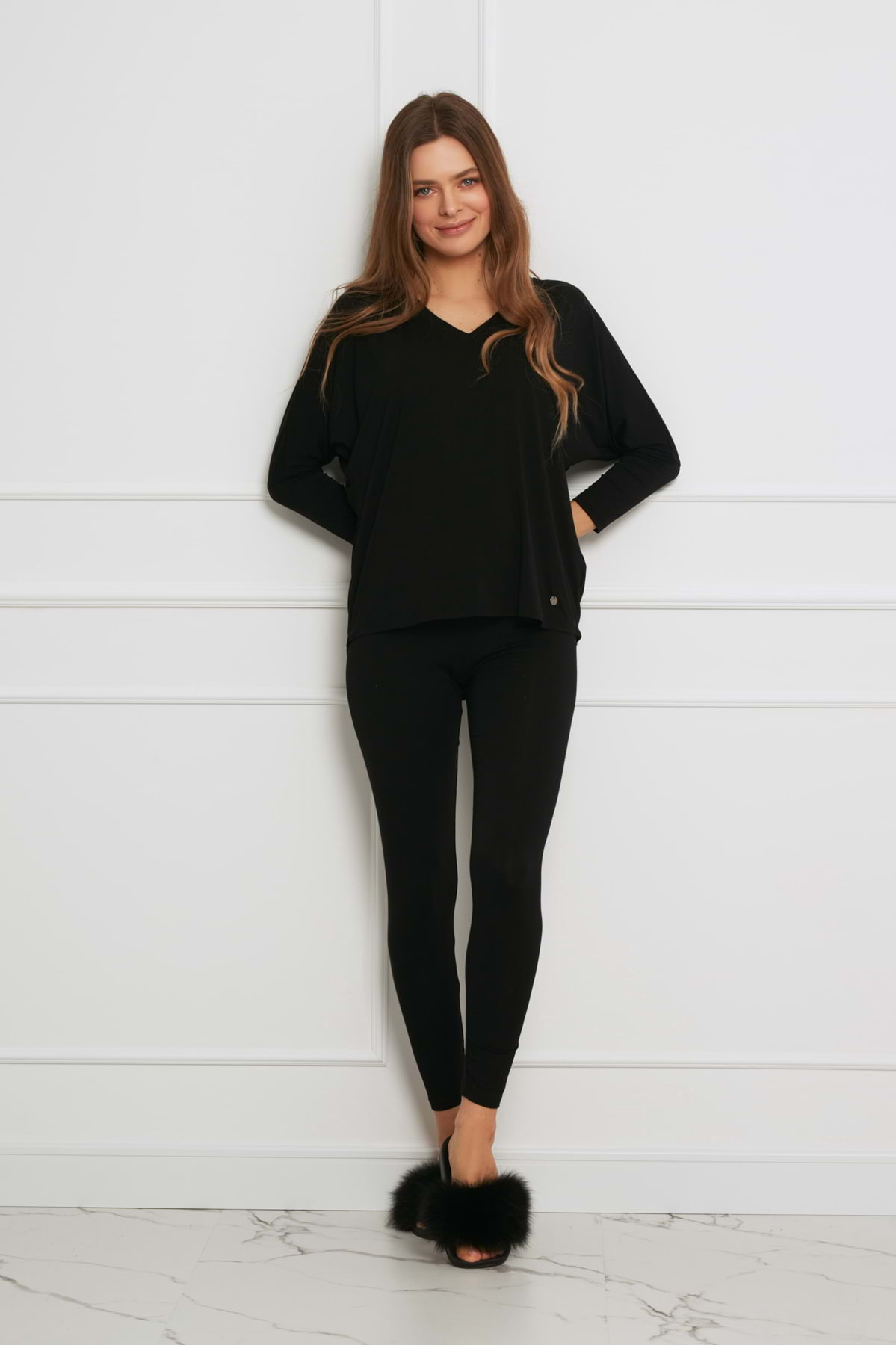 Kobieta z długimi brązowymi włosami nosi Loungewear wiskozowy komplet z legginsami - czarny, stojąc przy białej ścianie z listwami i marmurową podłogą, uśmiechając się z rękami za plecami.