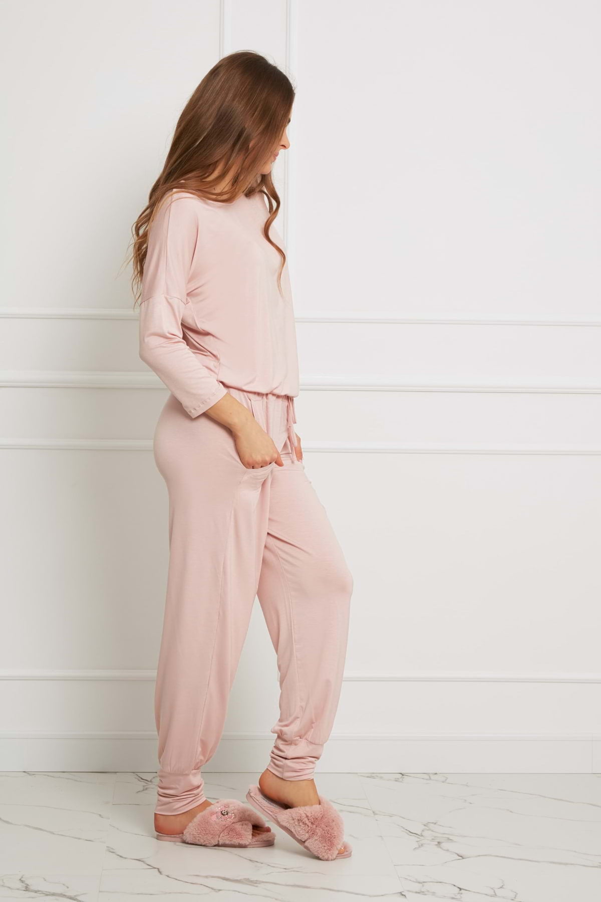 Kobieta z długimi brązowymi włosami stoi bokiem w minimalistycznym pokoju, ubrana w Loungewear długi komplet ze spodniami z kieszeniami - brudny róż i pasujące puszyste kapcie. W tle białe ściany wyłożone panelami i marmurowa podłoga.