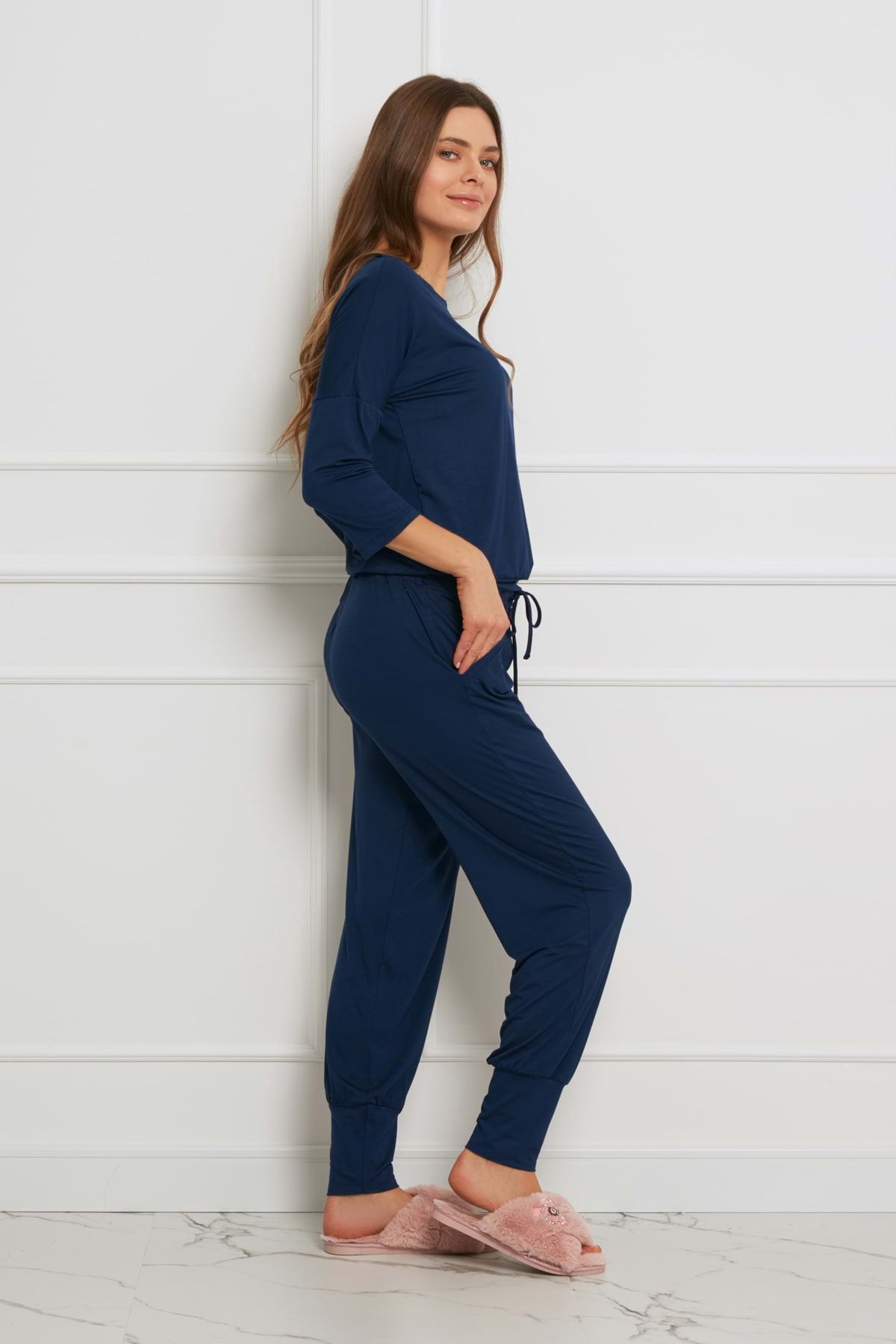 Kobieta stoi bokiem, uśmiechnięta, ubrana w granatowy Loungewear długi komplet ze spodniami z kieszeniami i jasnoróżowe rozmyte kapcie, na tle białej ściany i marmurowej podłogi.